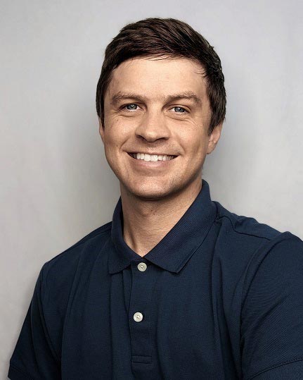 The image shows a man with short hair smiling at the camera. He has light skin and is wearing a blue polo shirt with a collar. The background is neutral, suggesting a professional setting.
