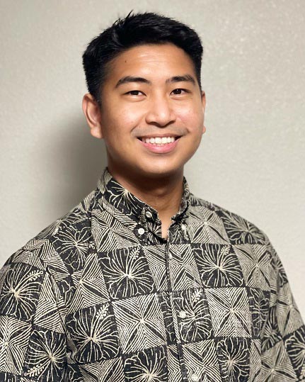 The image shows a smiling man wearing a patterned shirt standing against a neutral background.