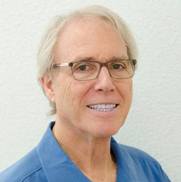 The image shows a man wearing glasses, with short hair, dressed in a blue shirt and smiling at the camera. He appears to be indoors, possibly in an office setting, against a white background.