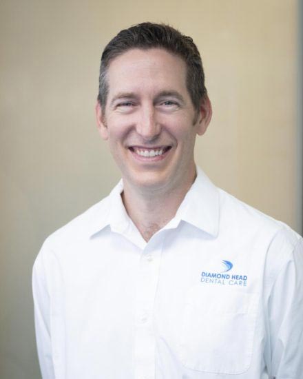 The image shows a man with a broad smile, wearing a white shirt with a logo on the left side, posing for a professional portrait.