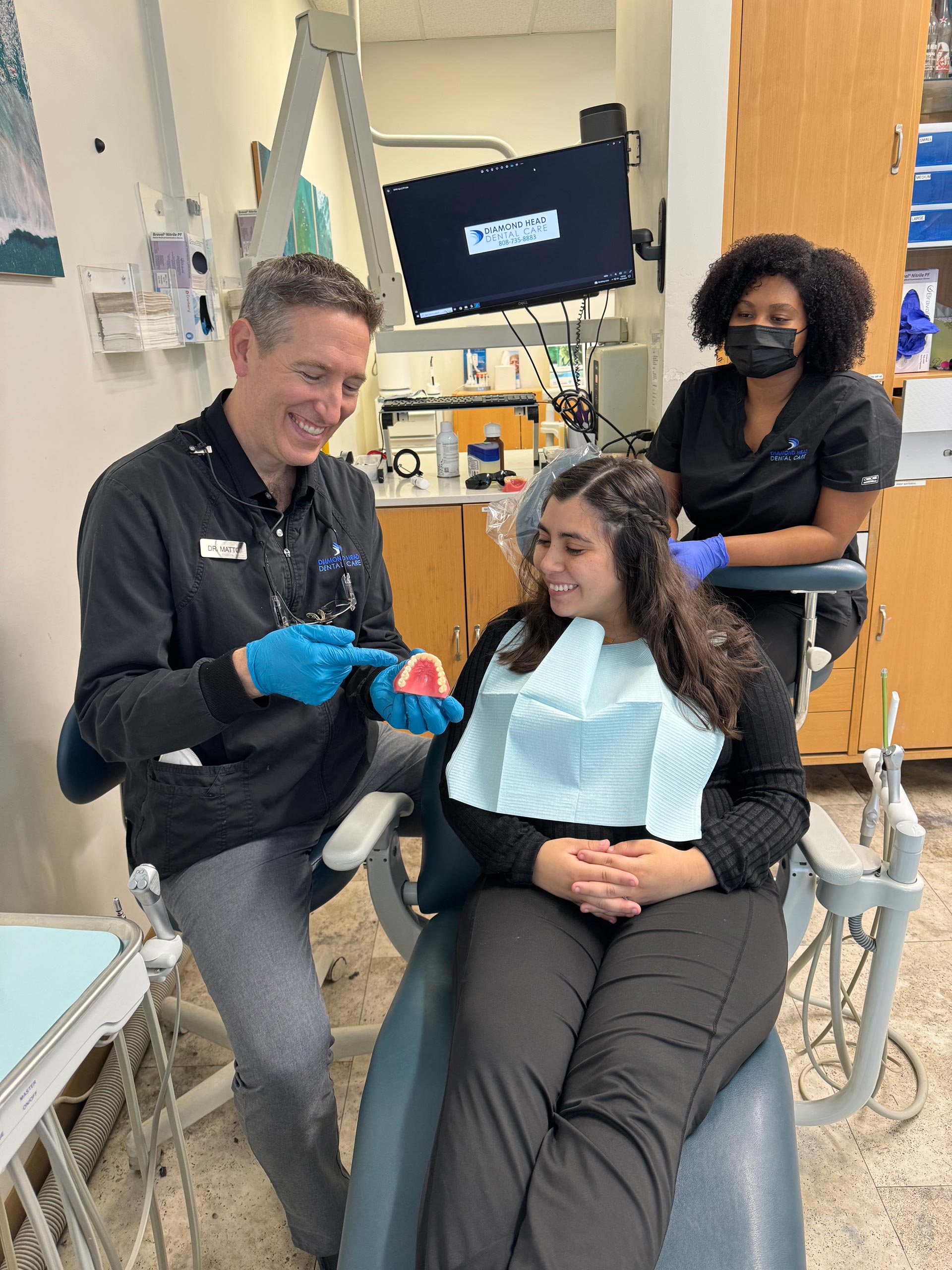 In the image, there is a dental professional holding a red object near a patient's mouth, both are inside a dental office setting with equipment visible in the background.