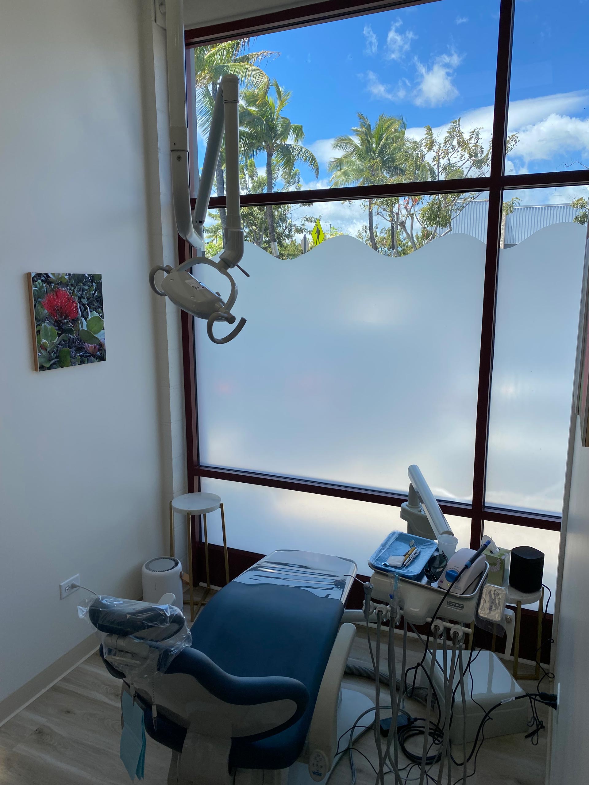 The image shows an interior view of a dental office with a large window allowing natural light to enter, displaying a blue sky outside. A dental chair is positioned under a window, and various dental equipment such as a dental mirror, lamp, and other instruments are visible on the countertop in front of it.