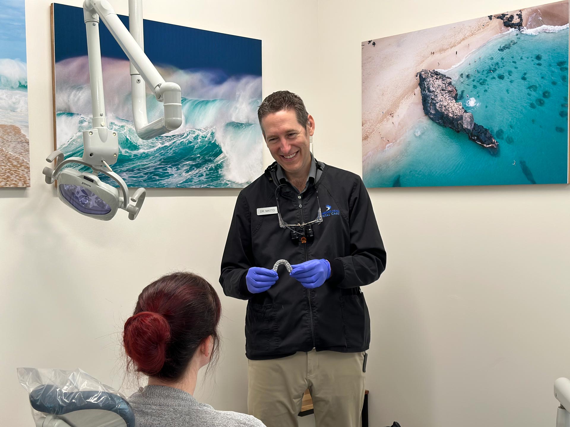 A dental professional, possibly an orthodontist, in a clinic setting, instructing a patient on their oral care routine, with a backdrop of ocean-themed artwork.