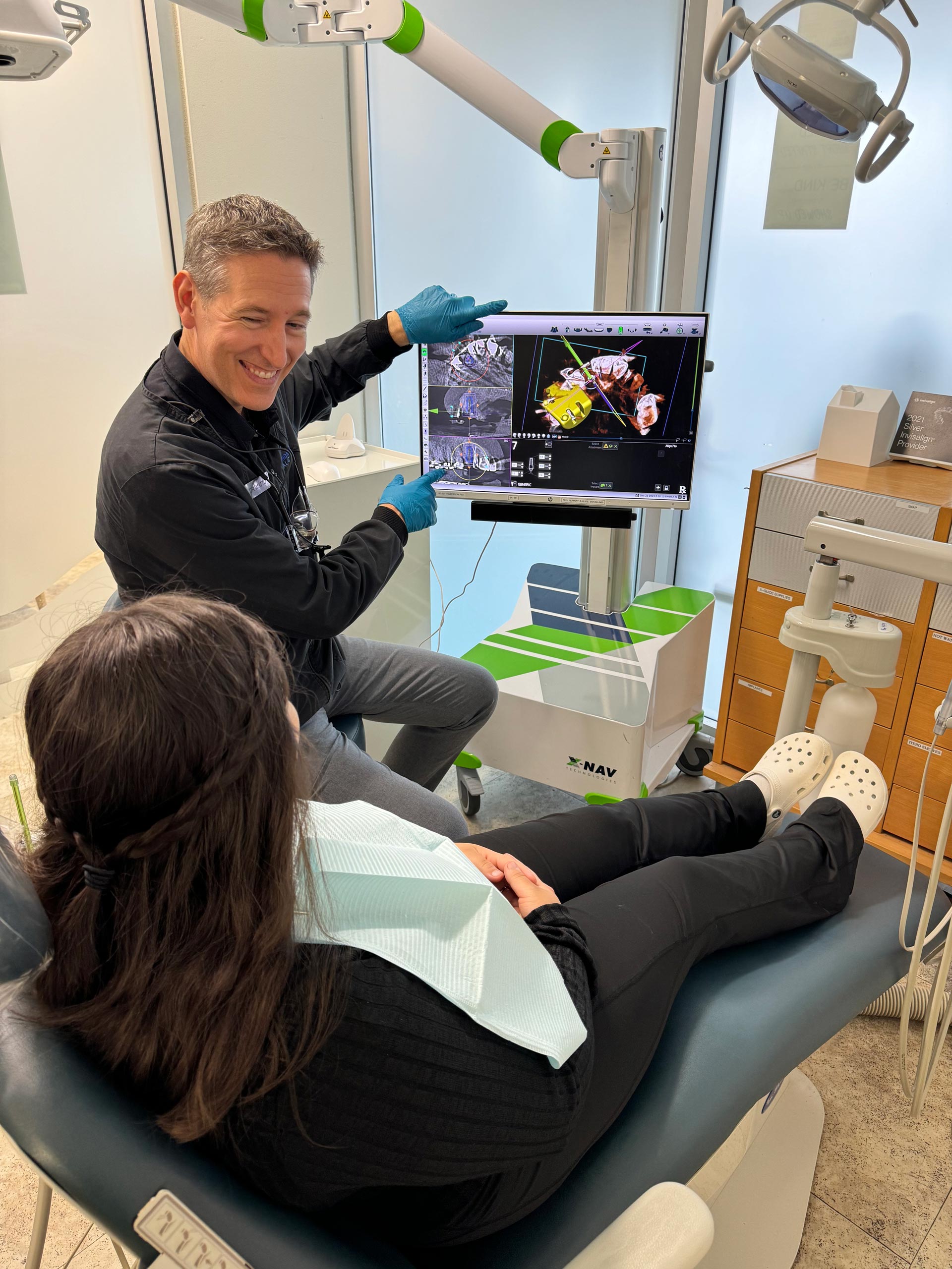 In the image, there are two people in a dental office setting. A man stands at a computer screen displaying an X-ray, while a woman sits in a dental chair with her mouth open, possibly receiving treatment.
