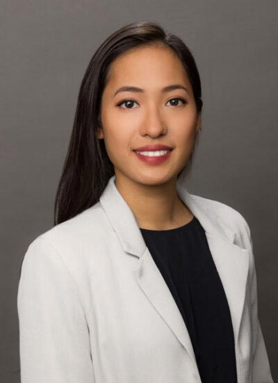 The image shows a person standing against a neutral background, smiling slightly at the camera. They have dark hair, are wearing makeup, and have a light-colored blazer over a black top.