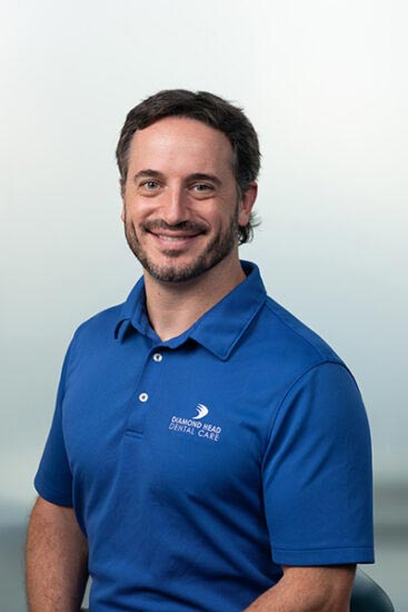 The image shows a man wearing a blue polo shirt, standing against a white background. He has short hair and is smiling towards the camera.