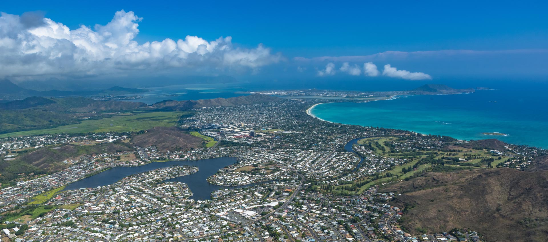 The image shows a scenic view of an island with a clear blue sky, lush green hills, a large body of water, and a town with buildings visible in the distance.