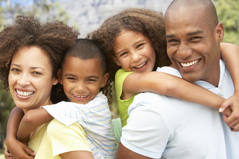 The image depicts a family of four with two adults and two children, smiling and posing for a photo outdoors during the daytime.