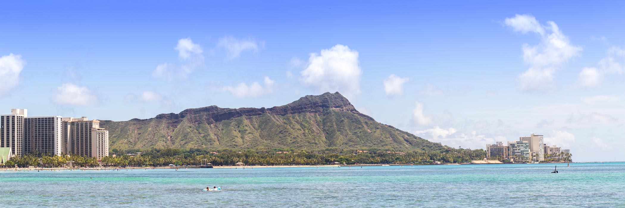 The image shows a panoramic view of a tropical landscape with an expansive body of water, a large island with buildings on the horizon, and clear skies.