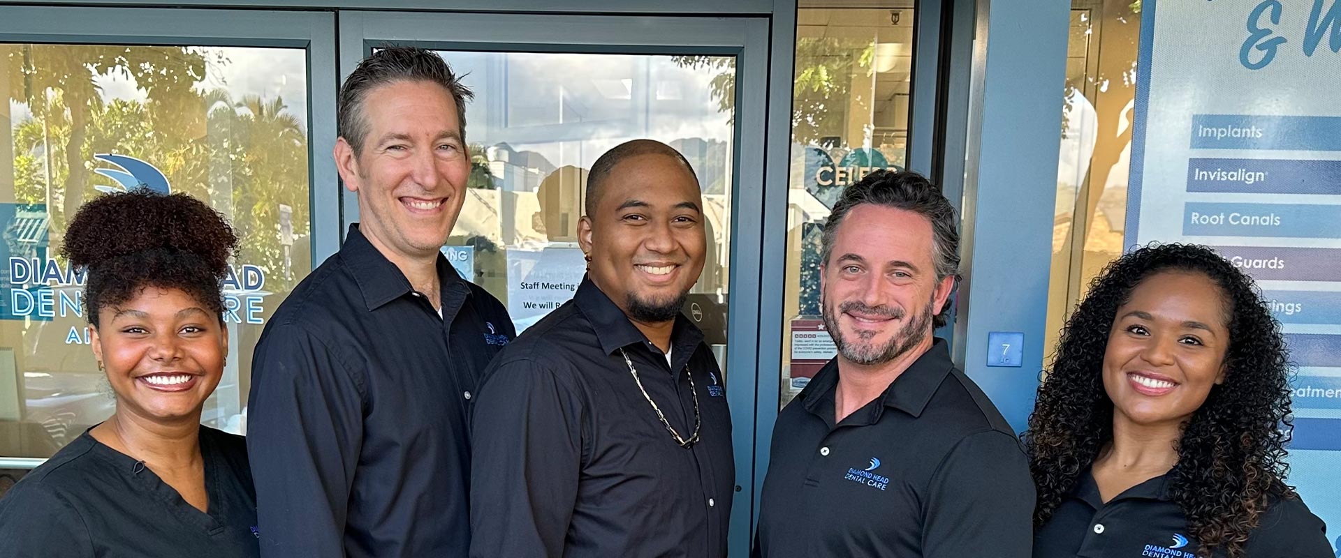 The image shows a group of people standing together, with some smiling and posing for the camera. They appear to be at an entrance to a building, as indicated by the signage behind them.