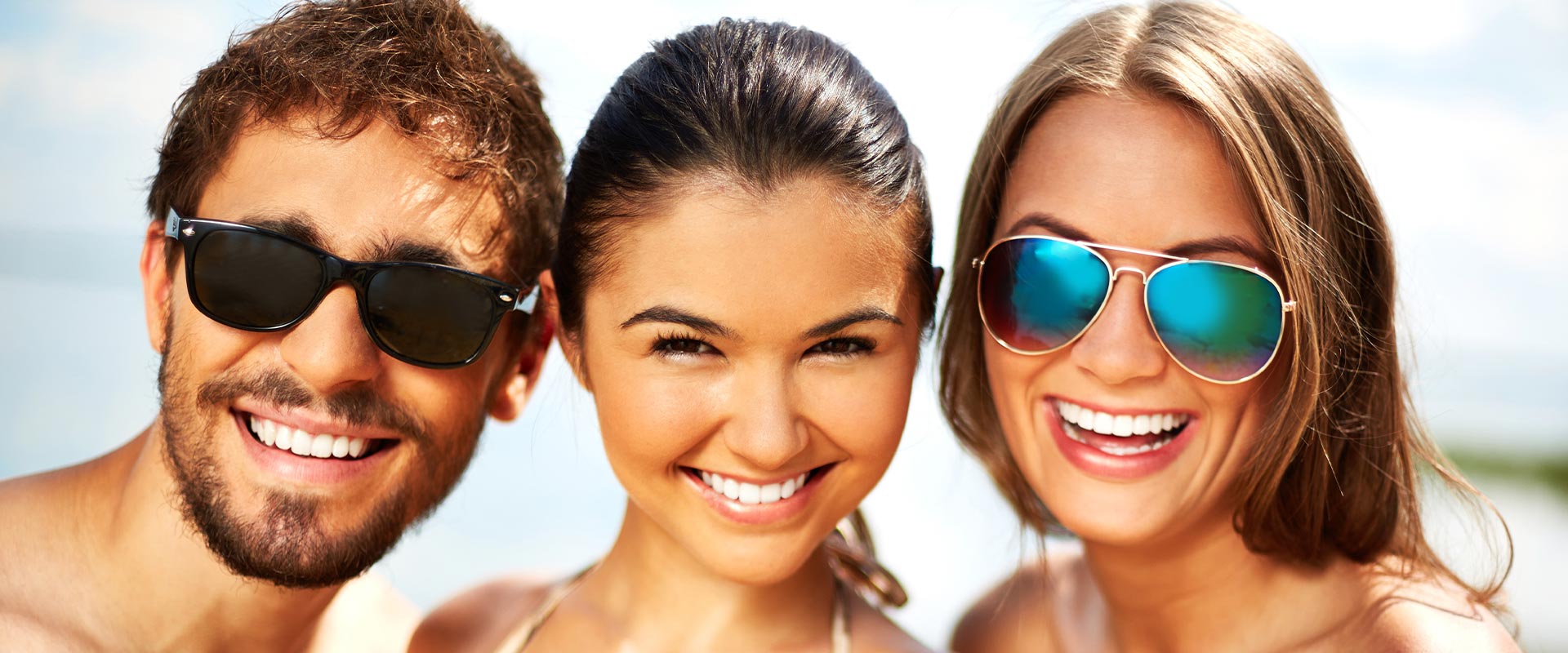 The image shows four people smiling at the camera with sunglasses on their heads  they appear to be enjoying a sunny day outdoors, likely at a beach or pool setting.