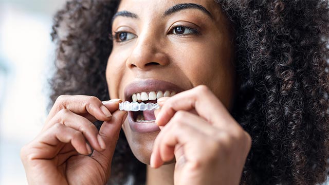 The image shows a person with dark hair wearing a face mask, brushing their teeth with a toothbrush.