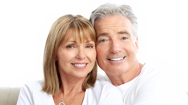 The image shows an older man and woman posing closely together, both smiling, with the man s arm around the woman s shoulder.
