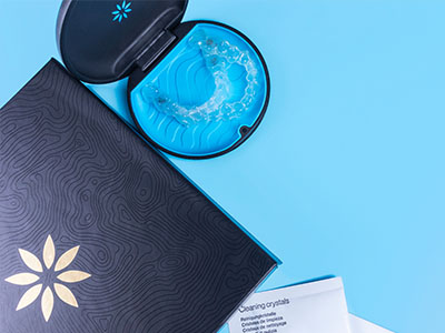 The image shows a cosmetic product with a blue lid placed on top of a dark-colored packaging box, which features a floral design and a white label with text and decorative elements.