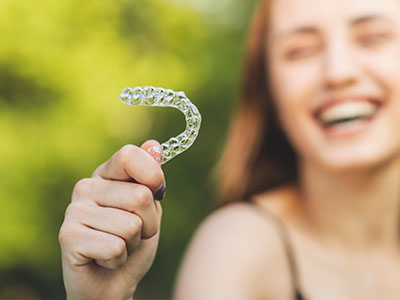 This is a color photograph featuring a smiling person holding up a clear plastic smiley face with a toothbrush-like shape, suggesting dental hygiene.