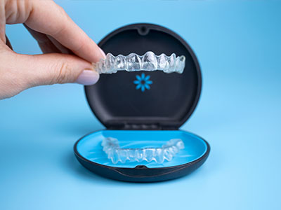 The image depicts a hand holding a clear plastic tray containing a set of transparent dental retainers, with the retainers partially inserted into an open blue case designed for storage.