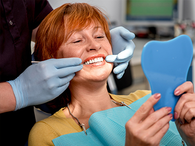 The image shows a person with red hair sitting in a dental chair, smiling at the camera while holding a blue toothbrush-shaped object, with a dental hygienist adjusting her smile by removing the device from her mouth using tweezers.
