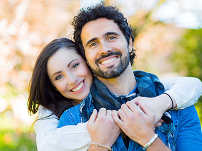 Man and woman hugging each other outdoors, with man wearing a blue shirt and woman in white, smiling and embracing.