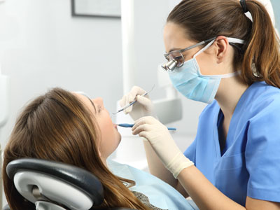 The image shows a dental hygienist performing a teeth cleaning procedure on a seated patient, with both individuals wearing personal protective equipment such as gloves and masks.