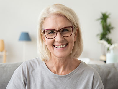 The image features a woman with blond hair smiling at the camera, wearing glasses and a grey top, sitting on a couch with a lamp visible behind her.