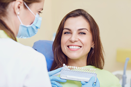 The image shows two individuals at a dental office one person is seated with a smile, wearing a surgical mask, and holding a dental device, while another person stands behind them, wearing gloves and a face mask, providing assistance.