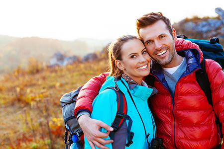 A man and woman are standing close together in an outdoor setting, smiling at each other, with the man wearing a backpack and both dressed in hiking gear.