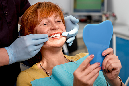 Woman sitting in dental chair receiving teeth cleaning, with dental hygienist holding blue toothbrush up to her mouth.