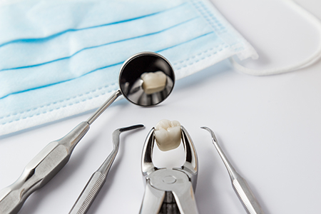 The image shows a dental hygiene setting with various dental tools including a toothbrush, dental pick, and dental mirror placed next to a blue surgical mask on a white surface.
