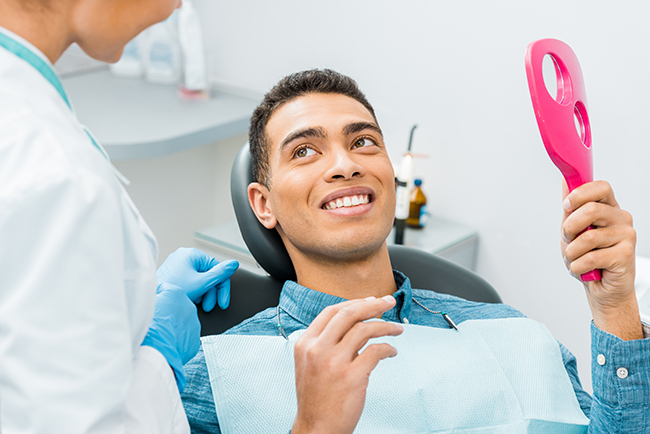 A man sitting in a dental chair with a smile on his face while holding up a pink object, possibly an oral hygiene tool, with a dental professional attending to him behind him.