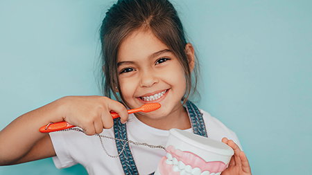 The image shows a young girl brushing her teeth with an electric toothbrush while holding a cup of toothpaste, smiling at the camera.