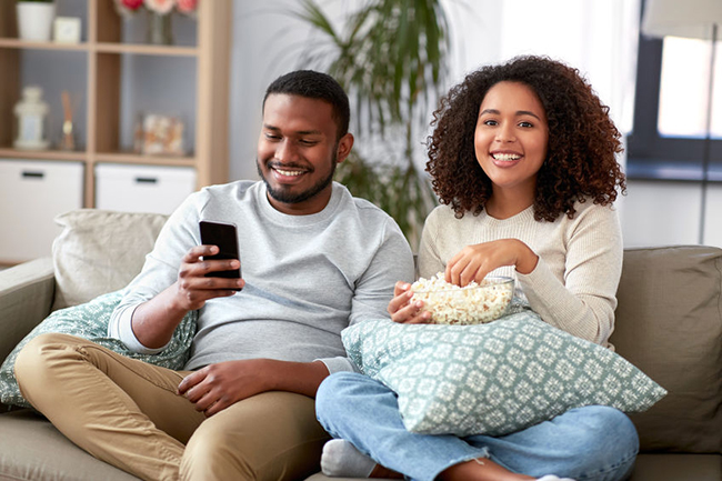 A man and woman are sitting on a couch, smiling and enjoying each other s company while watching something on a mobile device.