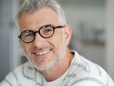 The image features a man with glasses smiling at the camera, appearing to be in his middle years, with graying hair and a beard, wearing a white shirt with a patterned collar.