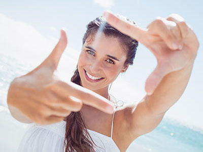 The image shows a woman taking a selfie with her hand up to the camera lens, smiling broadly at the camera, against a clear blue sky background with ocean waves visible in the distance.